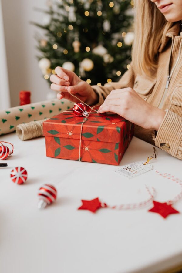 A woman wraps a festive Christmas gift with decorations on a table indoors.